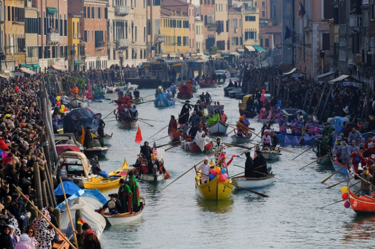 A flotilla of costumed rowers and decorated boats filling the canal between crowded banks at the water parade of The Venice Carnival, an engagement in the season calendar of The Mansa Court