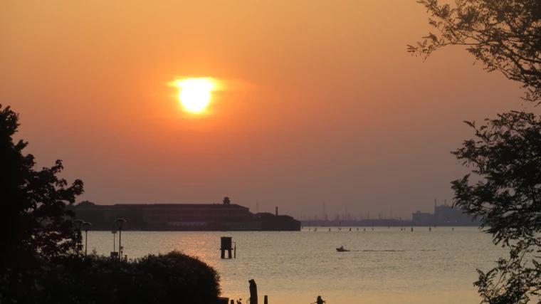An orange sun sets low over the Venetian lagoon, framed by dark trees, with a small boat crossing the water (2017).