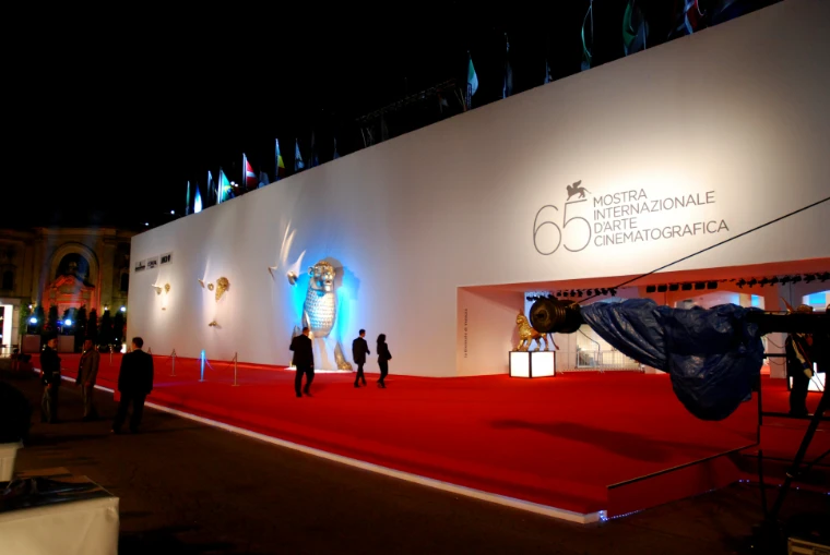 An empty red carpet runs at night along a white festival wall bearing the 65th Mostra logo and a gilded lion statue lit in blue (2008).