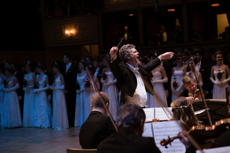 The conductor at full stretch on the podium with debutantes ranked in white behind him at The Vienna Opera Ball, an engagement in the season calendar of The Mansa Court