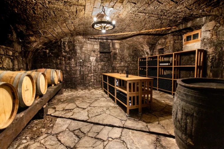 A vaulted stone cellar with oak barrels along one wall, wooden racks against the back and a hanging iron lamp above a flagstone floor, for The Vintners’ Table, New York in the season calendar of The Mansa Court