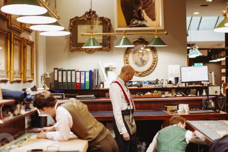 Tailors at work at their cutting tables in a busy workroom beneath green-shaded lamps, a gilt mirror on the wall, for The Wardrobe, New York in the season calendar of The Mansa Court