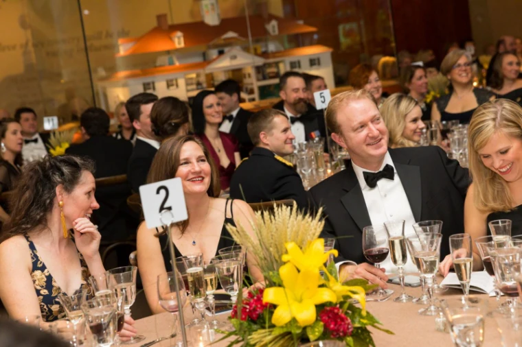 Round tables laid with candles and posies in a gilded crimson room before dinner at The Washington Birthday Ball, New York, an engagement in the season calendar of The Mansa Court
