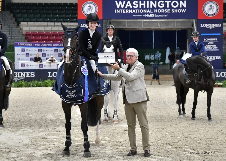 A rider on a dark horse in a blue Longines winner’s rug being handed a Longines presentation box in the arena, other riders mounted behind (2023).