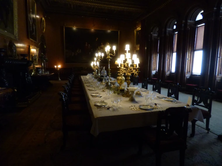 A long dining table laid with white linen, silver, glassware and lit gilt candelabra in a dark panelled dining room (2018).