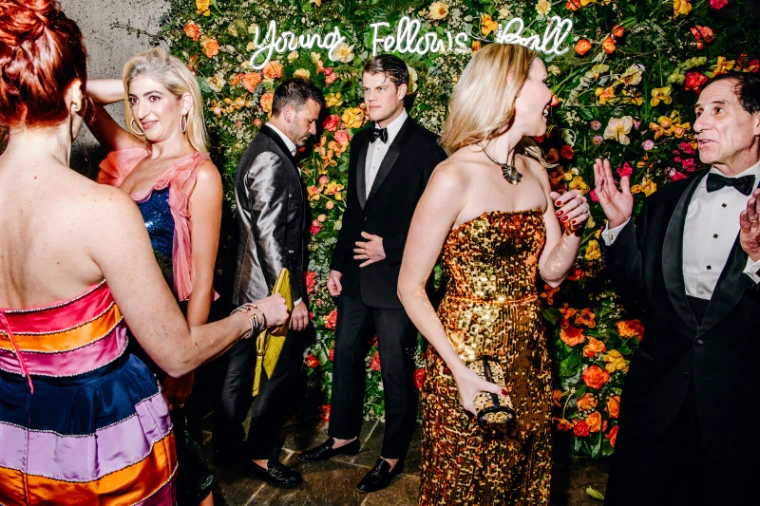 Guests in black tie before the flower wall and Young Fellows Ball sign at The Frick Collection, New York, an engagement in the season calendar of The Mansa Court