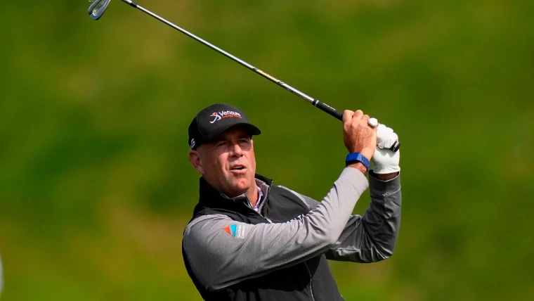 A golfer in a black cap following the flight of an iron shot at the top of his swing, a green background, for US PGA Championship, United States in the season calendar of The Mansa Court