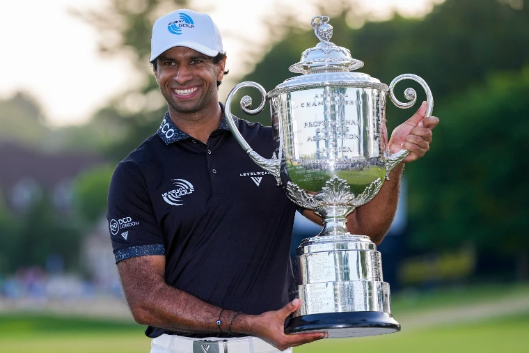 A smiling golfer in a black shirt and white cap holding the silver Wanamaker Trophy before trees and buildings, for US PGA Championship, United States in the season calendar of The Mansa Court
