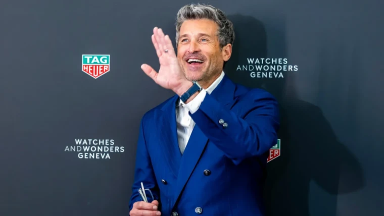 A guest in a blue double-breasted suit waving at the TAG Heuer photocall at Watches and Wonders Geneva, an early-April engagement in the season calendar of The Mansa Court